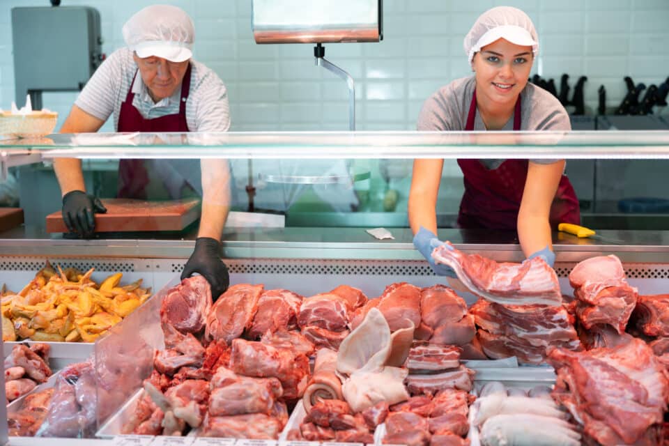 Young farmer woman in butcher shop takes raw pork ribs out of refrigerator