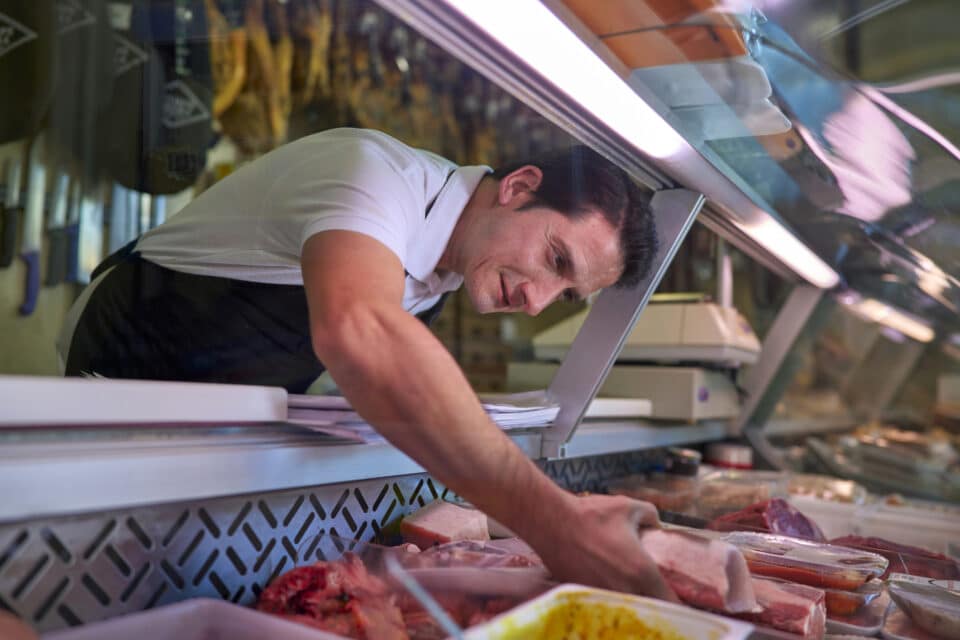 A Spanish butcher from a neighborhood butcher shop selects a meat from the refrigerated display to cut for his client