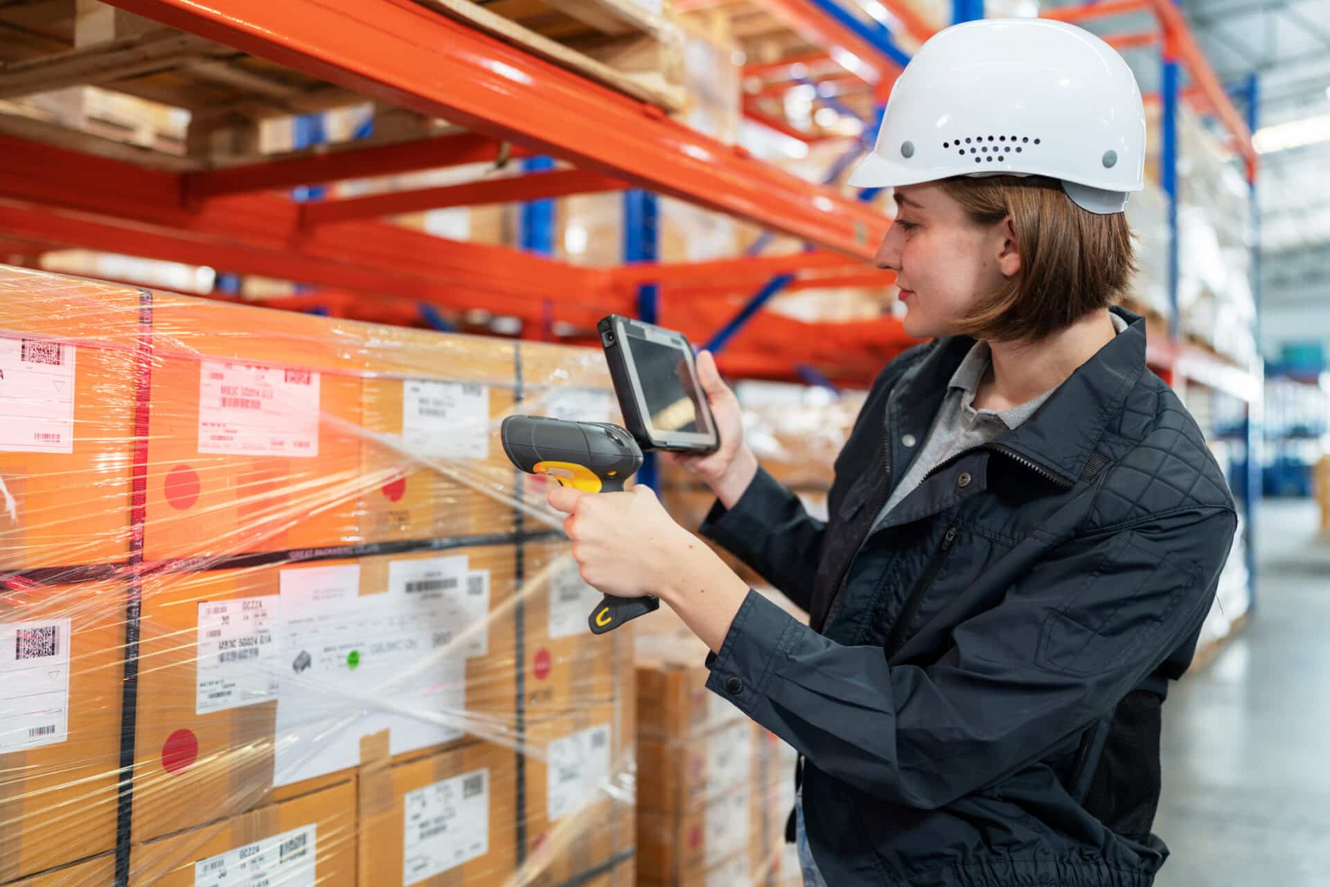 Implementing RFID tags help make delivery easier and growing up your business sustainably. Caucasian warehouse worker with safety helmet using a barcode reader to scan QR code on the box standing in the distribution warehouse. Looking at tablet