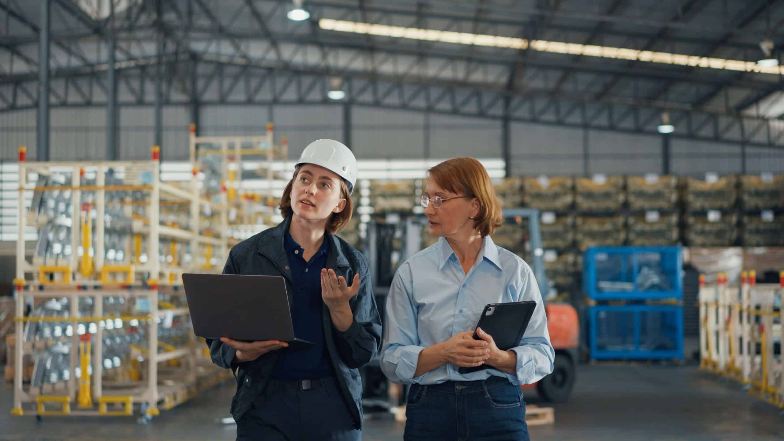 Eine junge kaukasische Mitarbeiterin und eine reife Managerin gehen durch das Lager eines Einzelhandelsunternehmens, überprüfen den Lagerbestand mit Tablet und Laptop und unterhalten sich dabei. Logistikbranche.