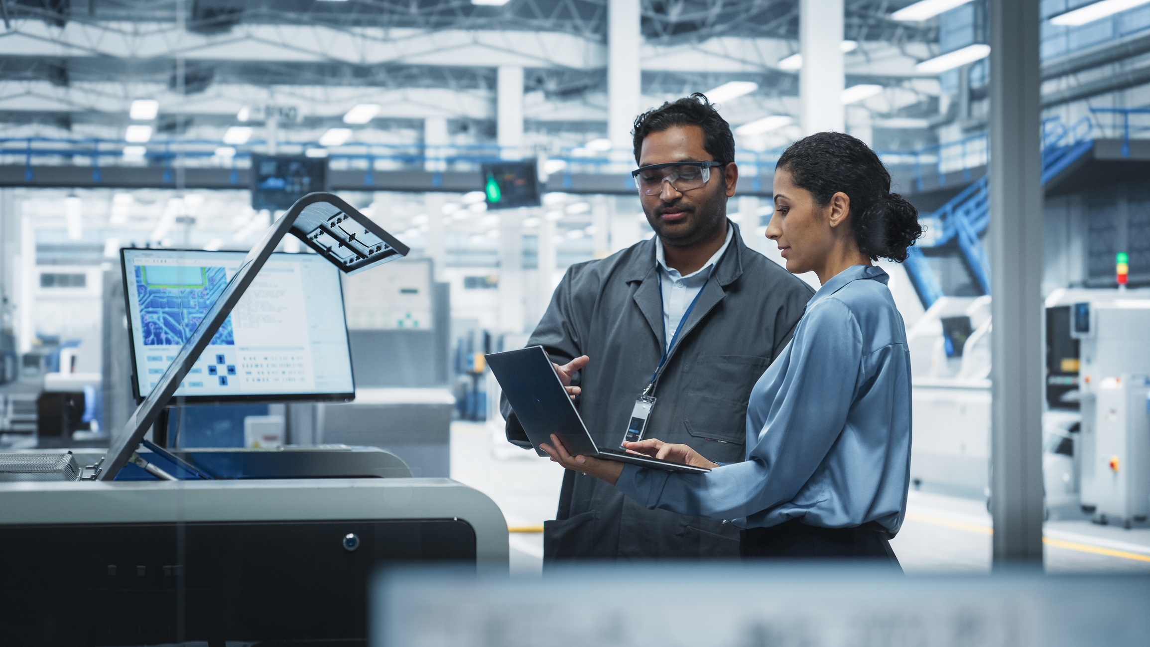 Indian Male Industrial Engineer And Hispanic Female Supervisor Using Laptop And Talking At Electronics Factory. Man Using Soldering Jet Printer, Explaining Process Behind Production Of Motherboards.
